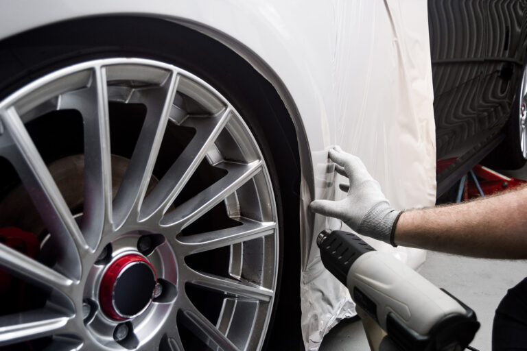 Ein Fachmann trägt mit weißen Handschuhen eine Schutzfolie auf den Kotflügel eines Autos auf. Im Vordergrund ist eine Heißluftpistole zu sehen, die für das Anbringen der Folie genutzt wird.