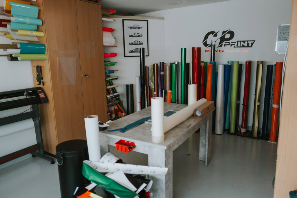 Kurs- und Arbeitsraum mit Werkbank, Plotter und vielen verschiedenen Folien in einer Reihe zum Aufbereiten von Autofolierungen