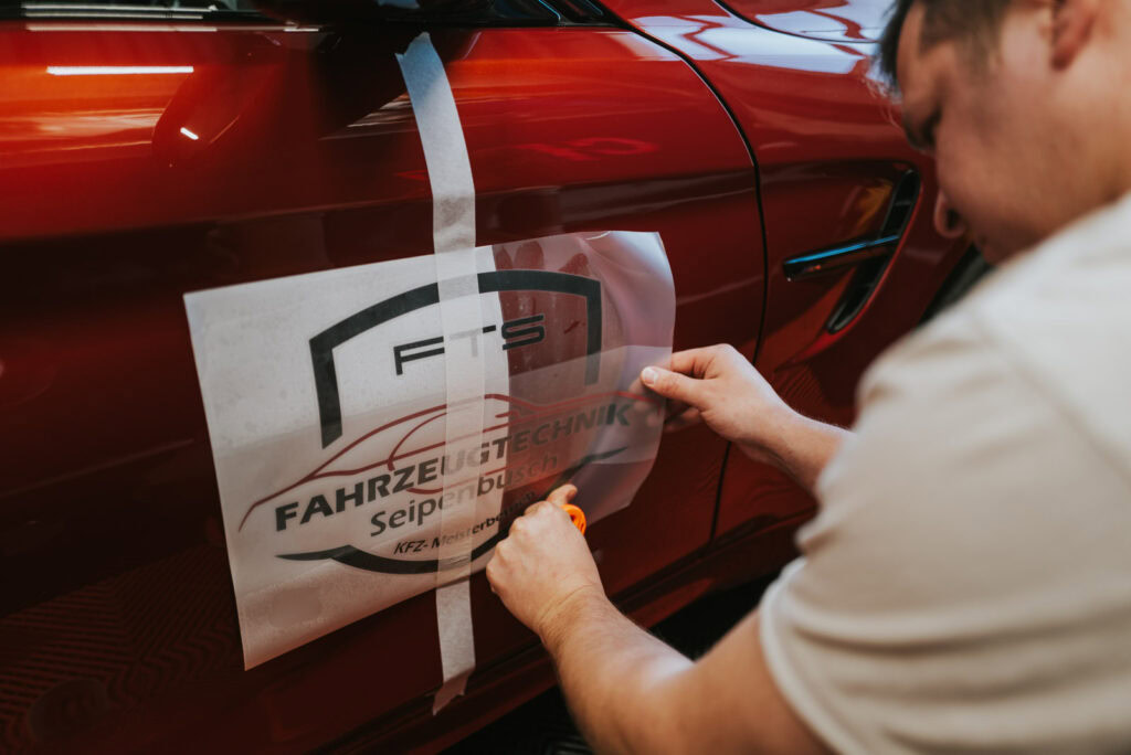 Kursteilnehmer kaschiert einen Schriftzug mit feinen Linien aus roter und schwarzer Folie auf sein Auto